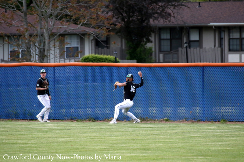 Two baseball players on a grassy field near a blue outfield fence; one pitcher in dark uniform throws the ball while a teammate stands nearby as a fielder watches.