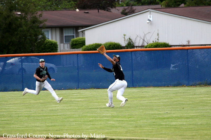 Two baseball players on a grassy field; one in black reaching up with a glove to catch a fly ball as a teammate runs nearby, blue fence in background.