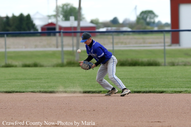 Youth baseball player in a blue uniform wrists a glove to field a ground ball on the infield dirt, with dust puffing up.