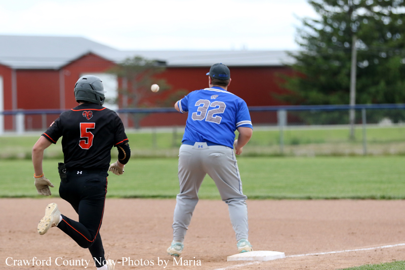 Two baseball players on a dirt infield near a base: a blue jersey runner (#32) stands on, while a black jersey fielder (#5) chases a ball midair with a red barn and outfield in the background.