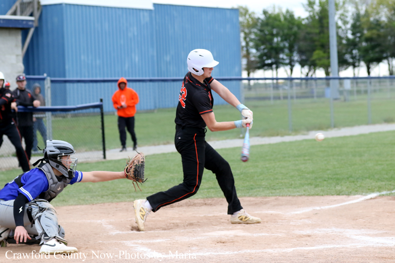 Baseball batter in a black uniform and white helmet swings at a pitch as the catcher in blue protective gear reaches to stop the ball near home plate on a sunny field.