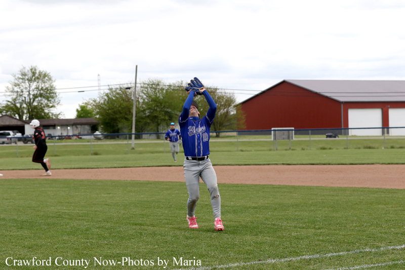 Blue-uniform baseball player reaches up to catch a fly ball on a grassy field during a game.