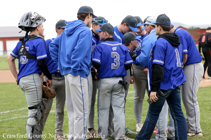 Youth baseball team in blue uniforms huddles on the field, players wearing helmets and gear, back numbers visible (e.g., 3 and 1).