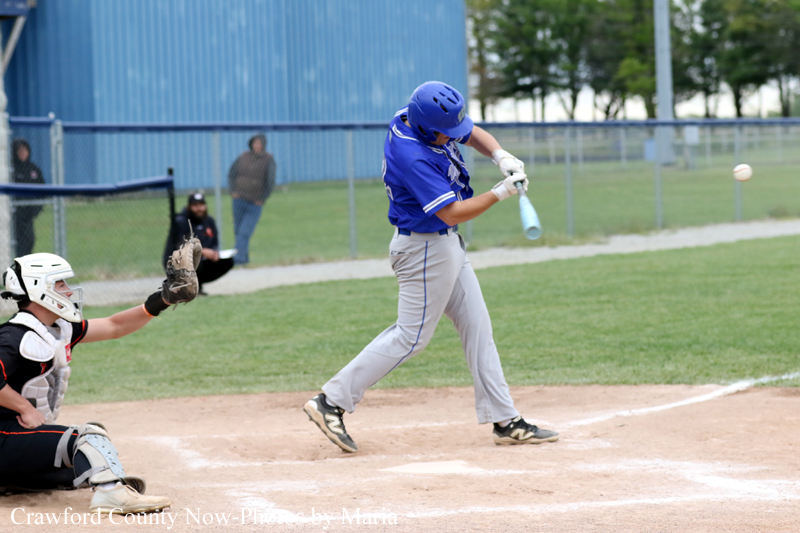Baseball batter in blue swings at a pitched ball as the catcher and umpire watch from behind the plate on a dirt infield.