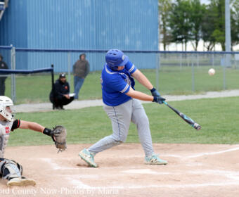 Blue-uniform baseball player swings at a pitched ball at home plate as a catcher wearing white and black gear crouches behind him on the dirt infield. Infield grass and a blue outfield wall are in the background.
