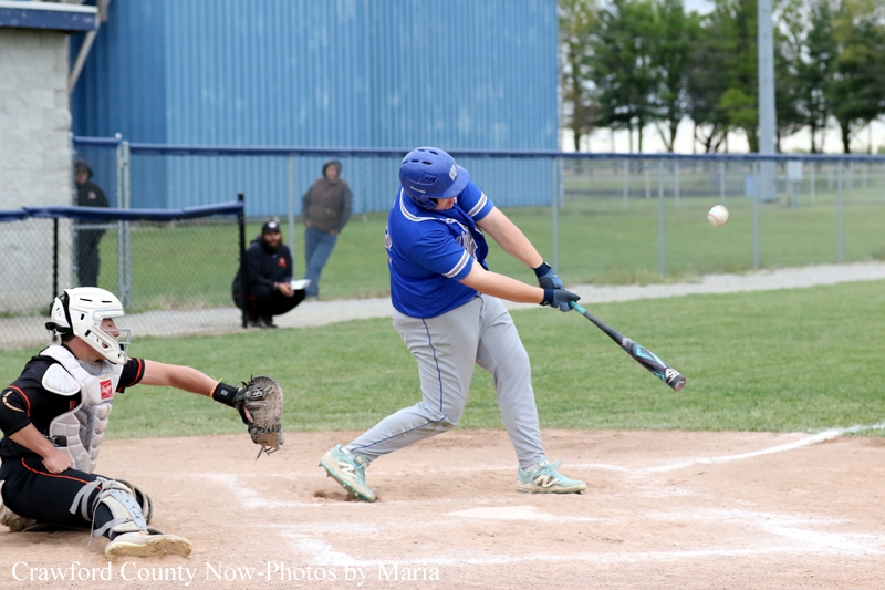 Blue-uniform baseball player swings at a pitched ball at home plate as a catcher wearing white and black gear crouches behind him on the dirt infield. Infield grass and a blue outfield wall are in the background.