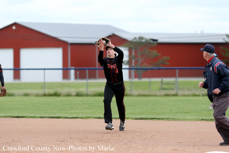 Baseball player in a black uniform reaches to catch a ball on the infield as an umpire watches nearby.