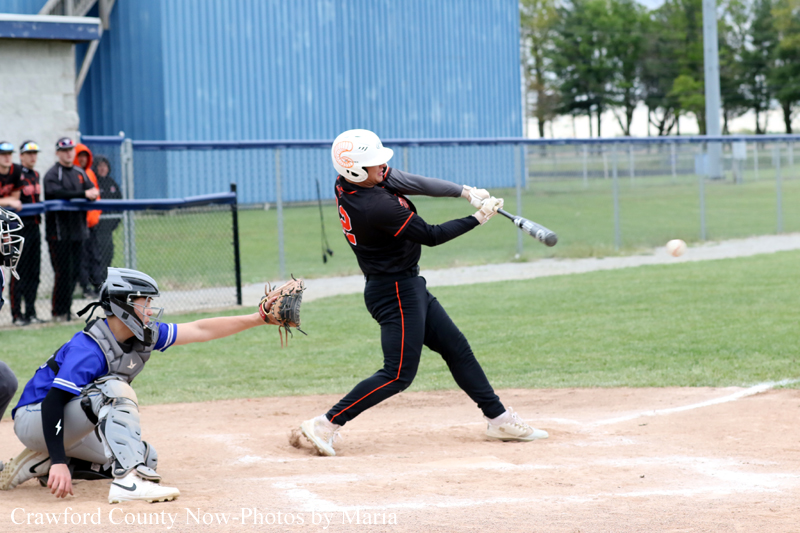 Baseball batter in a black and orange uniform swings at a pitch, with a blue-gray catcher crouched behind home plate on dirt.