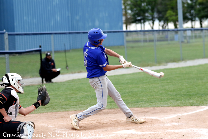 Baseball batter in a blue uniform and helmet swings at a pitch near home plate as the catcher crouches behind him in black gear.
