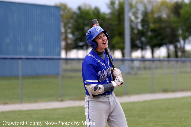 Baseball player in a blue uniform and helmet stands on the field, holding a bat over his shoulder and smiling.