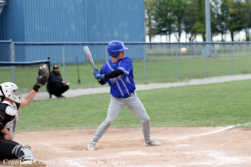 Baseball batter in a blue uniform at home plate, swinging as a pitched ball comes toward the plate; catcher crouches behind in gear.
