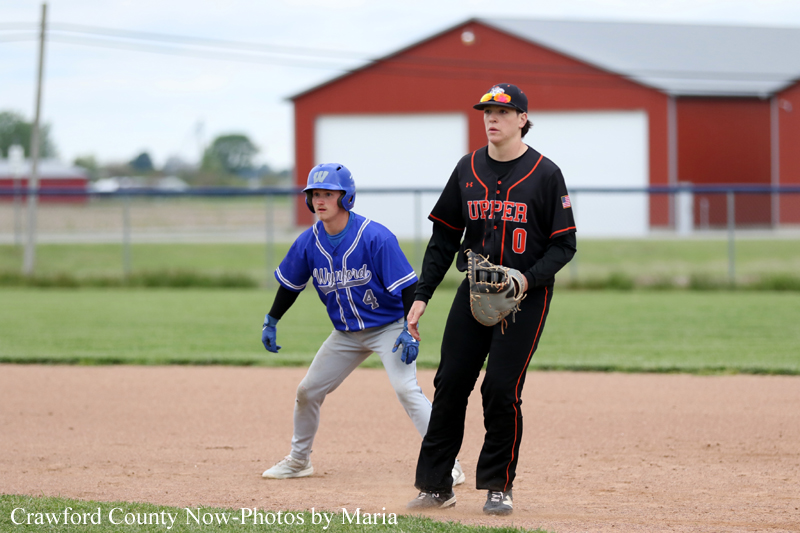 Two youth baseball players on the infield: a blue-uniform runner at first base and a black-uniform fielder watching the play with a glove, red building in the background.