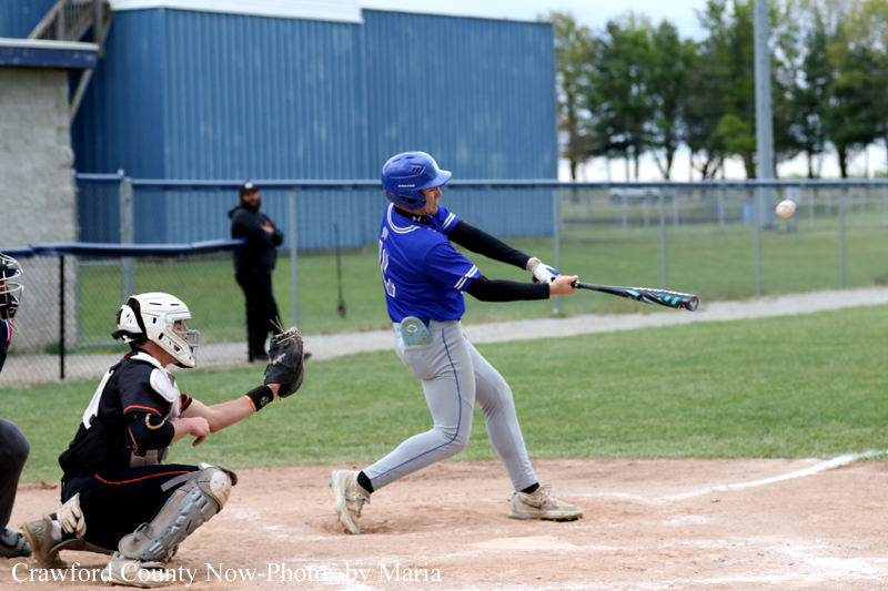 Baseball batter in blue jersey swings at a pitched ball as the catcher in black gear crouches behind him on the dirt field, with a blue outbuilding and fence in the background.