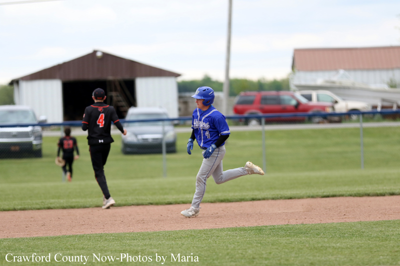 Boy in a blue baseball uniform runs on the dirt infield while a fielder in black chases behind, with rural buildings and parked cars in the background.