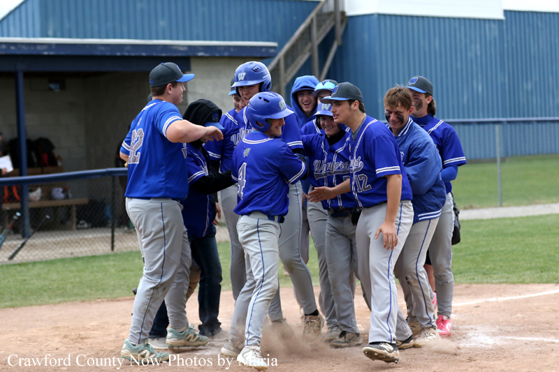 Group of baseball players in blue jerseys celebrate together on the infield near home plate, dust kicking up.