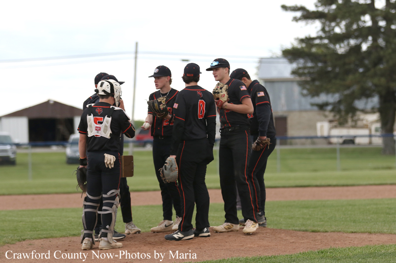 Six baseball players in black and red uniforms huddle on the infield dirt under an overcast sky, discussing strategy.
