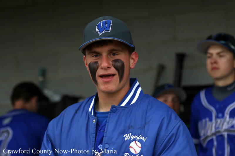 Baseball player in a blue jacket with a W cap, wearing black eye paint, standing in a dugout.
