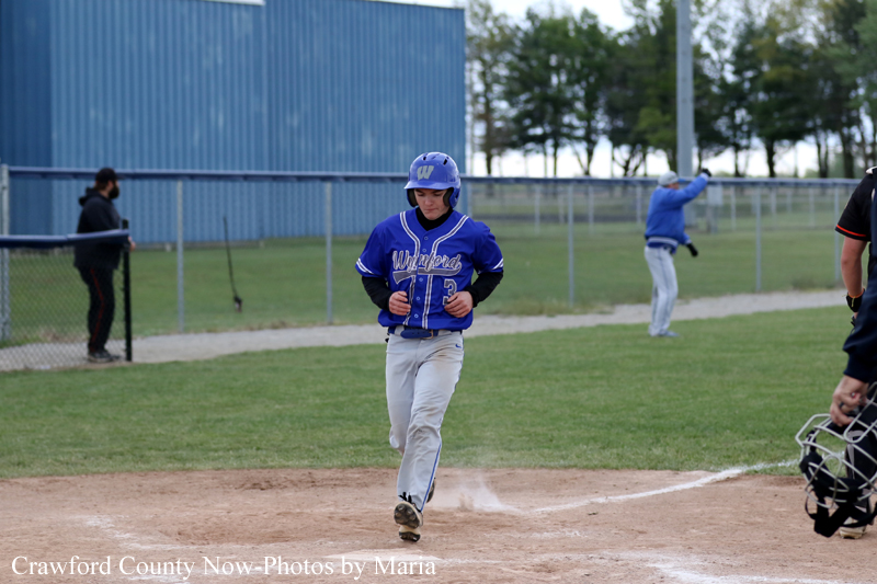 A baseball player in a blue uniform runs toward home plate as dust kicks up from the dirt. A catcher is visible on the right, and a coach/umpire stands in the background by the fence.