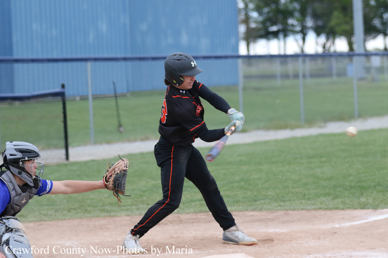 Young baseball batter in black uniform swings at a pitched ball at home plate as the catcher in gray gear reachs with a glove nearby.