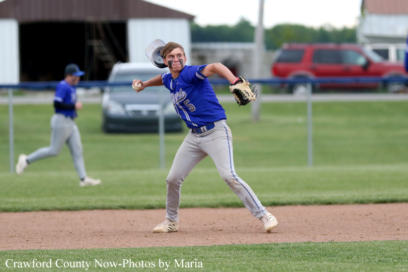 Youth baseball pitcher in a blue uniform delivering a pitch on the infield, with a teammate and field in the background.