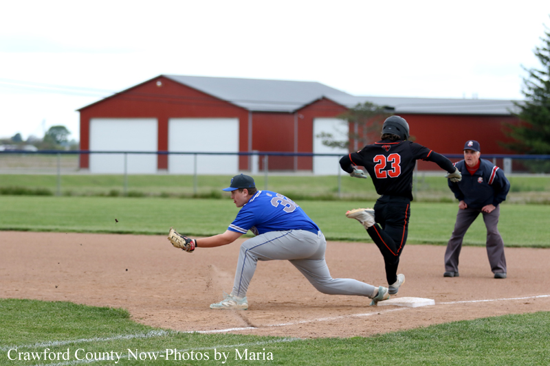 Blue-team infielder with glove reaches for the bag as a baserunner in black (#23) slides into base; umpire watches in the background on a baseball field.