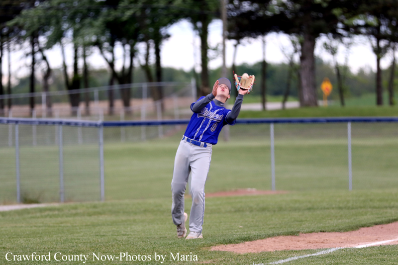 Baseball player in a blue uniform reaches upward with a glove to catch a ball along the infield dirt near the outfield fence.