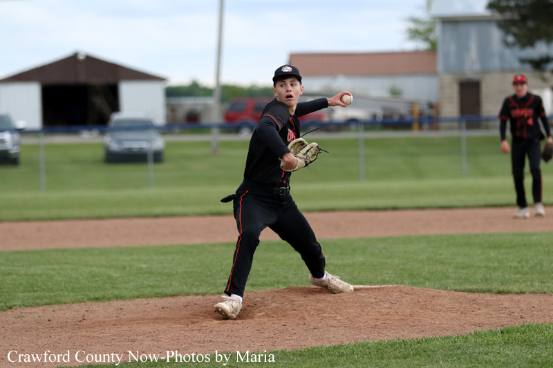 Baseball pitcher in a black uniform delivers a pitch from the mound, ball in hand, on a grassy field with a teammate in the outfield background.