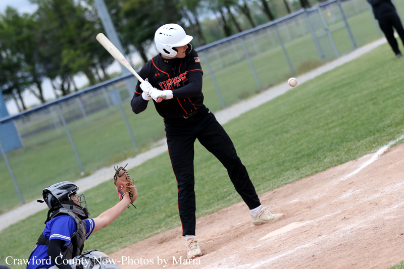 Baseball batter in a black uniform and white helmet swings at a pitch, with a blue-clad catcher crouched behind home plate and the ball in midair.