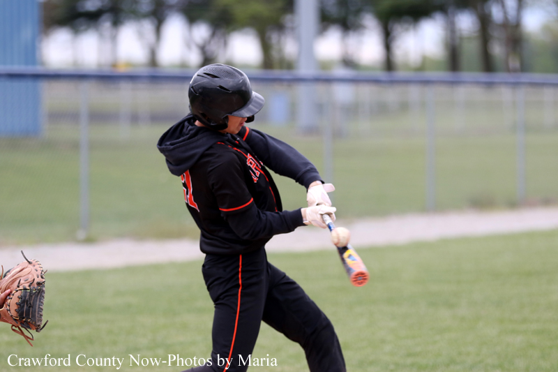 Young baseball player in a black and red uniform swings a bat during a game, wearing a helmet on a grassy field.