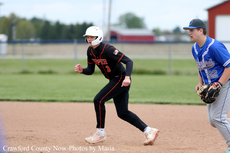 Young baseball player in a black and orange uniform and white helmet sprints on the dirt infield as a blue-uniform fielder watches from nearby with a glove in hand.
