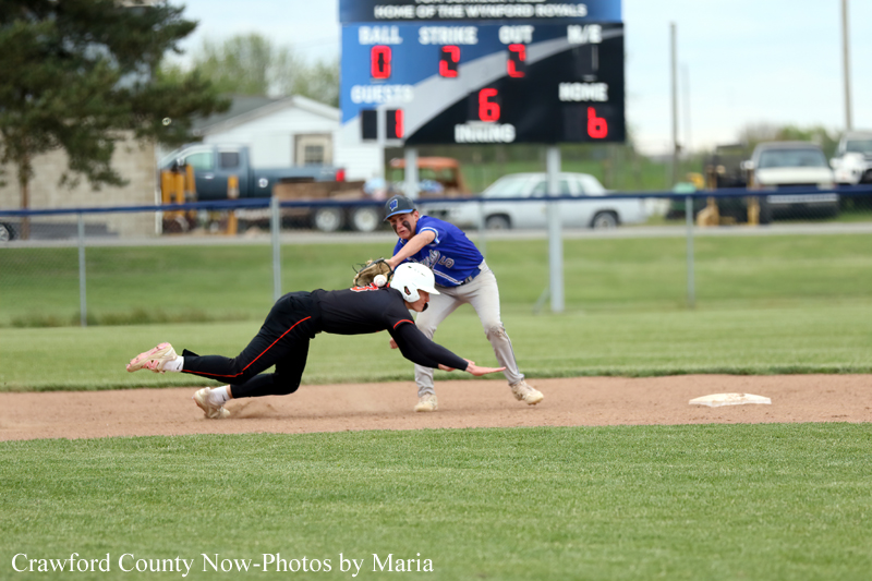 Baseball play: baserunner slides into second as the fielder reaches to tag; scoreboard and parked cars visible in the background.