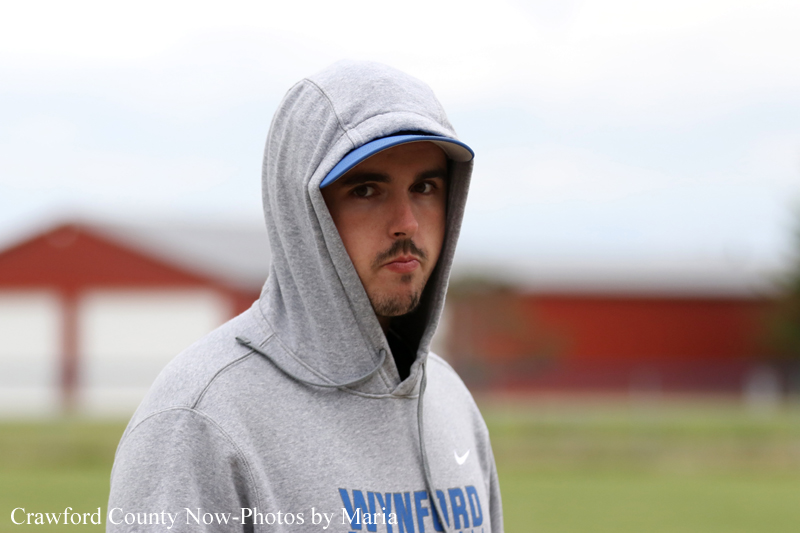 Portrait of a young man in a gray hoodie and blue cap, standing outdoors on a grassy field and looking at the camera.