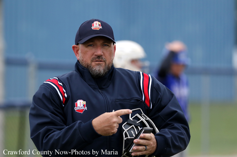 Baseball coach in a navy jacket and cap points while holding a catcher's mask on a field, with players in the background.