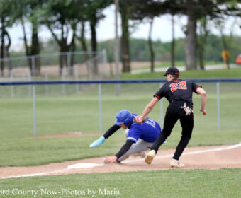 Baseball scene: runner in black (no. 22) slides into second as blue defender dives to tag him; dirt infield, grass outfield, fence and trees in background.