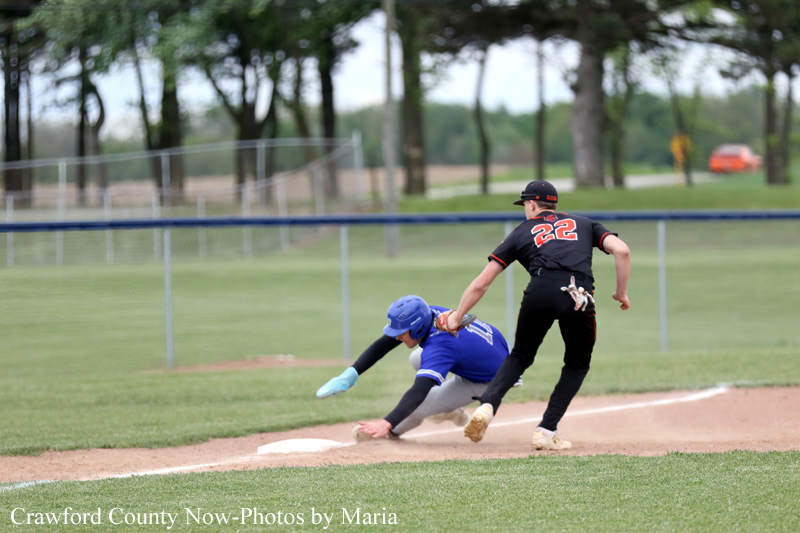 Baseball scene: runner in black (no. 22) slides into second as blue defender dives to tag him; dirt infield, grass outfield, fence and trees in background.