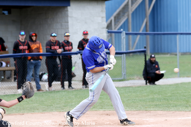 Baseball batter in a blue uniform swings a light blue bat as spectators watch from behind a chain-link fence behind the dugout area.