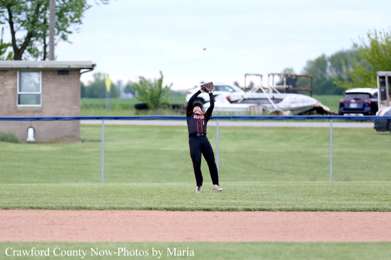 Baseball outfielder in a black and red uniform leaps to catch a fly ball near the outfield fence on a grassy field, with a building and boats visible in the background.