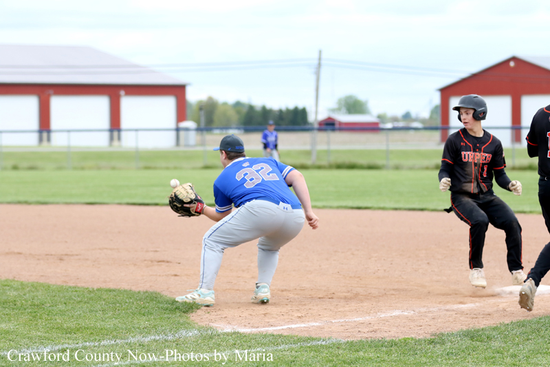 Baseball field action: a blue-team fielder with glove low to catch a ball near a base as black-uniform runners approach.