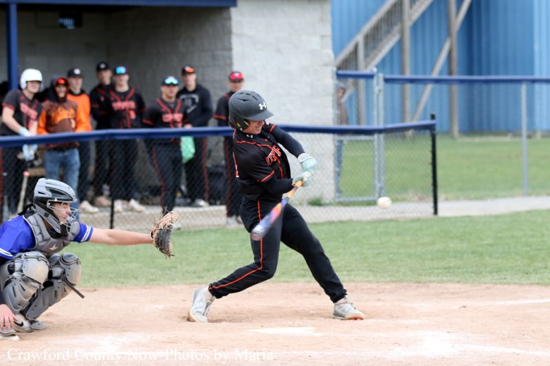 Batter in black uniform swings at a pitch as the catcher crouches behind home plate in protective gear by a dirt infield edge and players watch from the fence.”