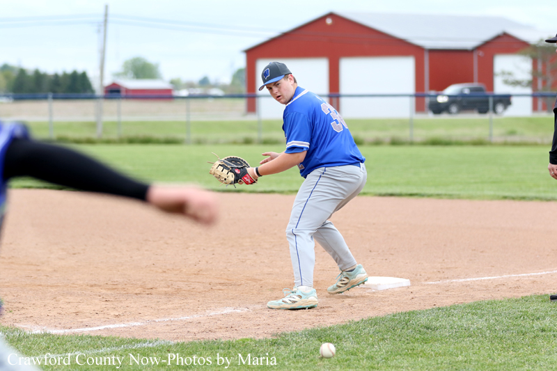 Baseball infielder in a blue uniform crouches at a base on a dirt infield, glove ready to field a ball, with a red barn and fence in the background.