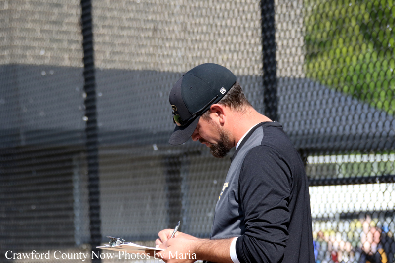 Bearded man in a dark cap and black long-sleeve shirt signs a clipboard near a chain-link backstop at a baseball field.