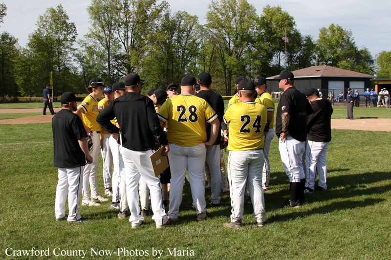 Baseball players in yellow jerseys form a huddle on a grassy field, numbers 28 and 24 visible on backs.