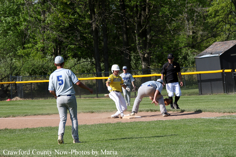 Youth baseball game on a sunny day: a runner in a yellow jersey slides into a base while a fielder in gray watches and the umpire stands nearby.