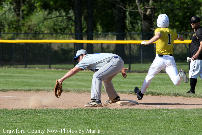 Baseball play at first base: a fielder in gray dives to scoop a ground ball as a dust cloud rises, while a runner in a yellow jersey (#1) runs past the bag.