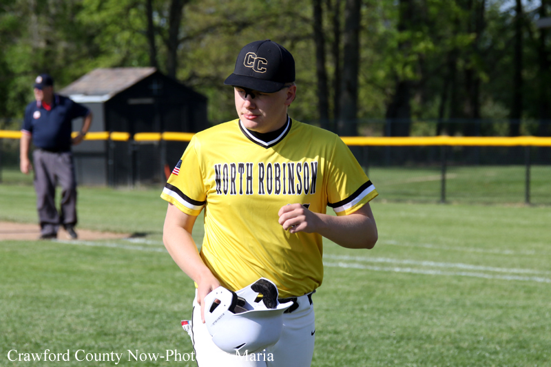 Young baseball player in a yellow North Robinson jersey walks off the field, helmet in hand.