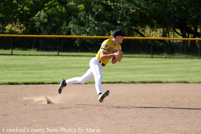 Baseball pitcher in a yellow uniform throws a pitch on the infield dirt with green grass and a fence in the background.