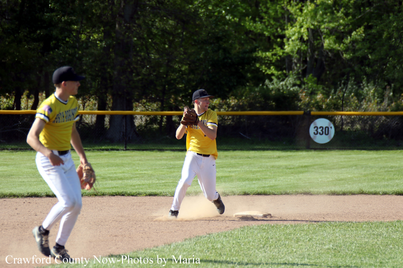 Two young baseball players in yellow jerseys on a sunlit dirt infield; one glove up ready to catch while the other approaches, dust rising from the ground behind them.