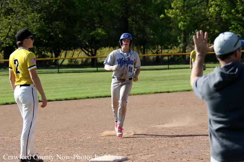 Baseball player in gray uniform with blue helmet runs on the dirt infield as a yellow-uniformed fielder watches nearby.