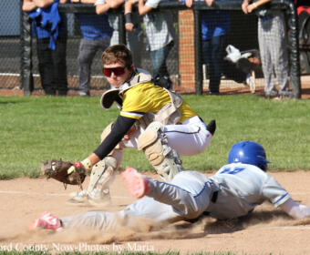 Catcher in yellow and white protective gear reaches for a sliding baserunner on the dirt infield, with spectators behind a chain-link fence watching the play.