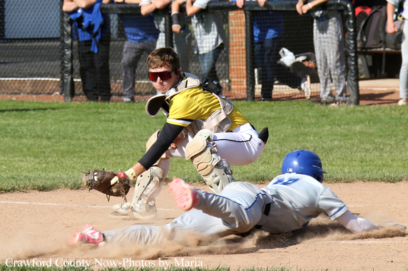Catcher in yellow and white protective gear reaches for a sliding baserunner on the dirt infield, with spectators behind a chain-link fence watching the play.